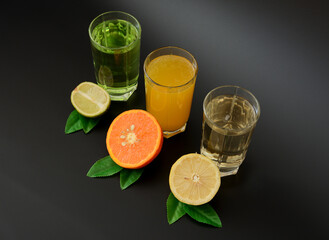 Three glasses with citrus juices, lemon, orange and lime decorated with leaves on a black background.