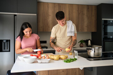Two young latin people laughing while peeling potatoes and cutting tomatoes to prepare a recipe at kitchen.