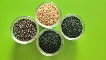 Nigella sativa (kalonji), black mustard, black sesame and white sesame seeds each displayed in separate bowls against an attractive green background