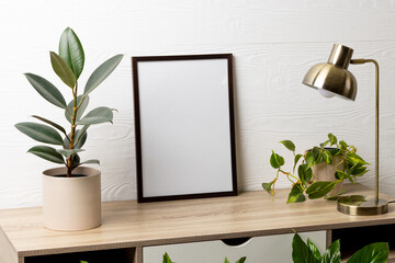 Black empty frame with copy space, lamp and plants in pots on table against white wall