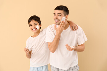Father and his little son applying shaving foam onto faces against beige background