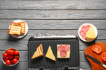 Composition with modern electric grill, delicious sandwiches and ingredients on grey wooden background