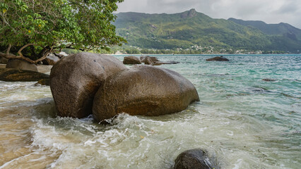 Large granite boulders are scattered in the surf of the ocean. Turquoise waves foam on the sand....