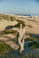 a woman in a light flying dress walks on the sand in sunglasses during sunset lighting