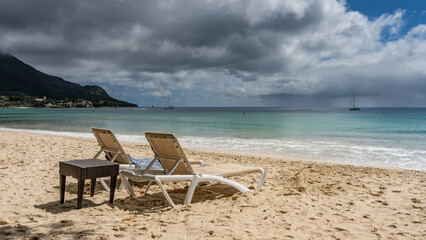 Two sun loungers and a wicker table stand side by side on the sandy beach. Ahead is a turquoise ocean, foam of waves. Silhouettes of yachts on the horizon. Seychelles. Mahe.