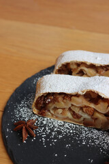 Traditional Apple strudel with cinnamon and powdered sugar cut in slices on a black slate stone plate on wooden table