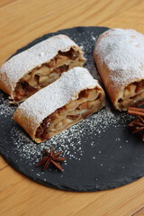 Traditional Apple strudel with cinnamon and powdered sugar cut in slices on a black slate stone plate on wooden table