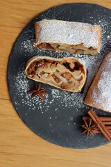 Traditional Apple strudel with cinnamon and powdered sugar cut in slices on a black slate stone plate on wooden table