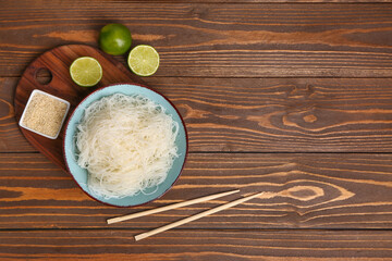 Bowl with tasty rice noodles, chopsticks, sesame seeds and lime on brown wooden table