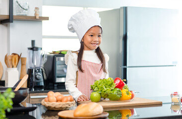 Millennial Asian young little cute girl chef daughter with white tall cook hat and apron standing smiling holding wooden spoon and fork posing ready for cooking vegetables at counter in home kitchen