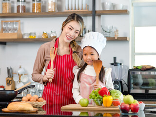 Asian young little cute girl chef daughter wears white tall cook hat and apron standing smiling posing together with mother holding red sweet pepper ready for cooking vegetables salad in home kitchen