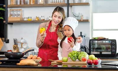 Asian young little cute girl chef daughter wears white tall cook hat and apron standing smiling posing together with mother holding red sweet pepper ready for cooking vegetables salad in home kitchen