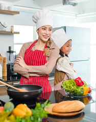 Asian young cheerful female mother and little cute girl daughter wearing white tall cook hat and apron standing crossed arms smiling posing holding thumbs up together ready for cooking in kitchen