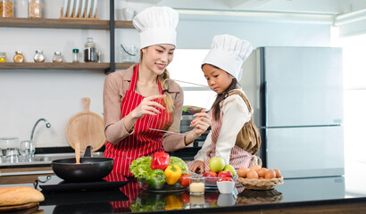 Asian young female chef housewife mother wears white tall cook hat and apron showing mockup glass touchscreen tablet computer screen to little cute girl chef learning cooking online at home kitchen