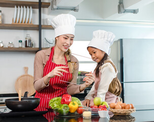 Asian young female chef housewife mother wears white tall cook hat and apron showing mockup glass touchscreen tablet computer screen to little cute girl chef learning cooking online at home kitchen