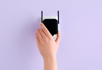 Woman plugging black WiFi repeater in electric socket on lilac wall