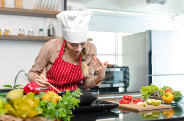 Millennial Asian young cheerful female housewife chef wearing white tall cook hat and apron standing smiling waving hand smelling tasty delicious meal while cooking in pan in full decorated kitchen