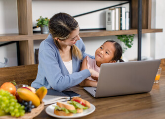 Asian young female mother and little cute girl daughter sitting smiling learning studying doing homework online via laptop notebook computer drinking orange juice eating sandwich together at home