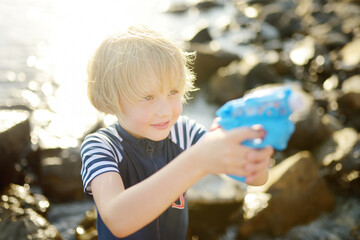 Cute little boy playing with water pistol on the seashore on a hot day. The child having fun with water gun outdoors during the summer holidays.