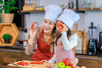 Asian young female chef housewife mother and little cute girl daughter wears white tall cook hat and apron smiling holding posing looking through sliced red sweet pepper with knife at home kitchen