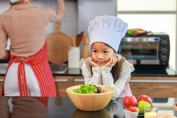 Millennial Asian young little cute girl chef daughter with white tall cook hat and apron standing smiling posing ready for cooking vegetables at counter in home kitchen.