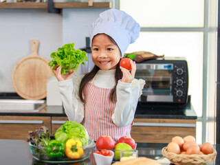 Millennial Asian young little cute girl chef daughter with white tall cook hat and apron standing smiling posing ready for cooking vegetables at counter in home kitchen.