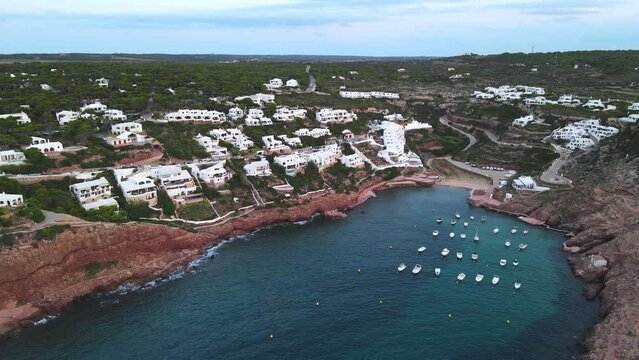 Luxury real estate on the side of cliff top in Menorca Spain with million dollar views. Drone zooms in to show ocean front properties with sailing yachts floating outside their back door.