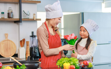 Asian young cheerful female chef housewife mother and little girl daughter wears white tall cook hat and apron standing smiling holding tulip flower bouquet together in kitchen preparing food meal