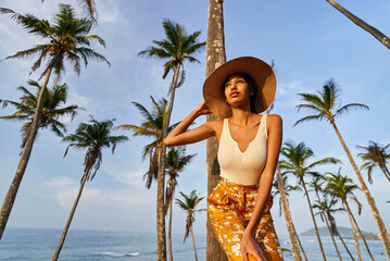 Young african female model posing in colorful clothes at tropical location at sunrise. Black woman against exotic scenery at dawn. Multiracial dark-skinned model poses in front of palm trees at sunset