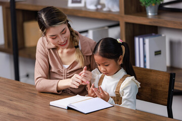 Asian young female housewife mother tutor teacher sitting smiling on table in living room at home teaching little cute kindergarten preschool girl daughter writing on book doing homework after school