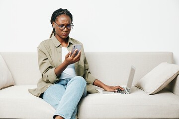 African American woman business freelancer working sitting on the couch at home in a laptop and phone, business calls and correspondence sadness and anger, home clothes and eyeglasses, light interior