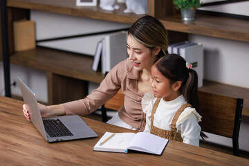 Asian young female housewife mother tutor teacher sitting smiling on table in living room using notebook computer pointing teaching little cute kindergarten preschool girl daughter doing homework