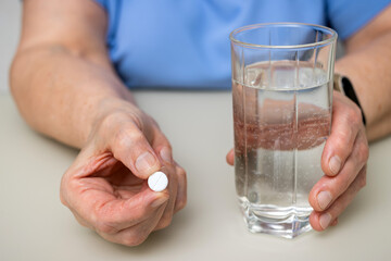 Senior woman with wrinkled old hands at the table holding one round medical tablet, painkiller or pill for treatment and water glass. Healthcare and medicine concept