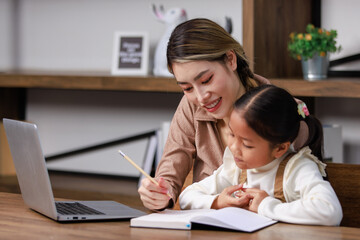 Asian young female housewife mother tutor teacher sitting smiling on table in living room using notebook computer pointing teaching little cute kindergarten preschool girl daughter doing homework