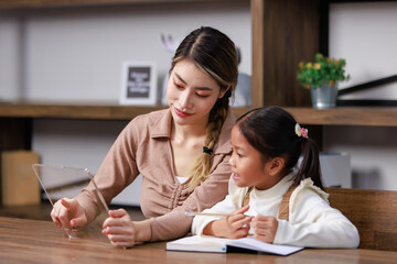 Fototapeta premium Asian young female housewife mother tutor teacher sitting smiling on table in living room holding tablet computer pointing teaching little cute kindergarten preschool girl daughter doing homework