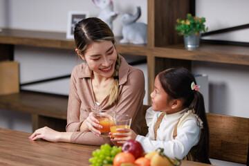 Asian young female housewife mother sitting smiling on table near bookshelf in living room at home with little girl kid daughter holding healthy organic orange juice glasses cheers drinking together
