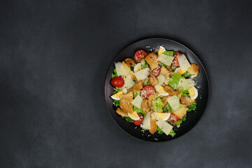 Top view of a Caesar Salad with chicken, lettuce leaves, cherry tomatoes, grated parmesan in a black plate against a Black Background - obrazy, fototapety, plakaty