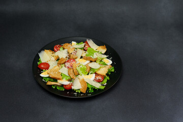 Caesar Salad with chicken, lettuce leaves, cherry tomatoes, grated parmesan in a black plate against a Black Background
