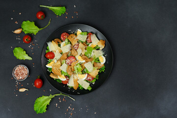 Top view of a Caesar Salad with chicken, lettuce leaves, cherry tomatoes, grated parmesan in a black plate against a Black Background