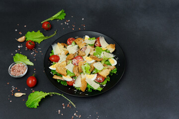 Top view of a Caesar Salad with chicken, lettuce leaves, cherry tomatoes, grated parmesan in a black plate against a Black Background