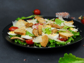 Close-up of a Caesar Salad with chicken, lettuce leaves, cherry tomatoes, grated parmesan in a black plate against a Black Background
