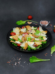 Caesar Salad with chicken, lettuce leaves, cherry tomatoes, grated parmesan in a black plate against a Black Background
