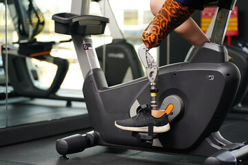 Young female with one prosthetic leg with exercising with a spinning bike in the gym to practice joint use with the other normal leg to be flexible, Concept of living a woman's life with a prosthetic