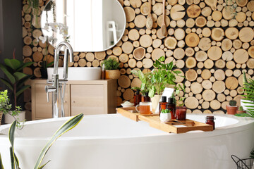 Stand with cosmetic products, cup, tablet computer and houseplants on bathtub in room