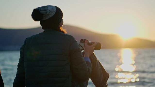A woman professional photographer taking picture of a sunset with a DSLR camera while sitting by the sea