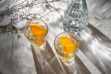 Glasses with orange infused water and bottle on beige table with aesthetic sunlight shadows,...