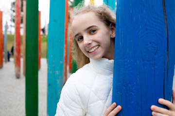 Portrait of a carefree teenage girl on the background of a blue wooden column. A cheerful blonde walks in a city park. Happiness and a smile on the face of a schoolgirl. Copy the space bar and empty.