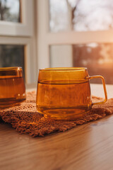 Two orange transparent glass mugs with hot tea with yellow reflection on a wooden table in the kitchen in sunlight. Top view