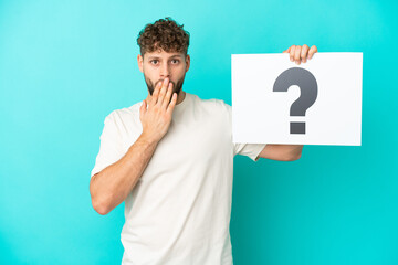 Young handsome caucasian man isolated on blue background holding a placard with question mark symbol with surprised expression