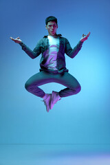 Fototapeta na wymiar Relaxation, calming down. Young emotive man in casual clothes jumping in yoga pose against blue background in neon light. Concept of human emotions, youth, fashion, lifestyle, feelings